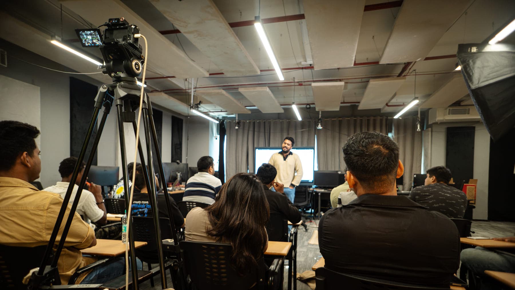 A GenC live class: the instructor at the front, a camera on a tripod recording, students at their desks